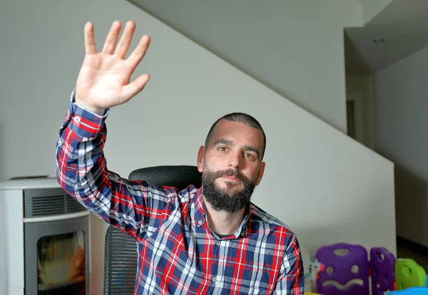 Young man saying hello to camera in a video conference from home. Baby playpen in the background. Work from home concept