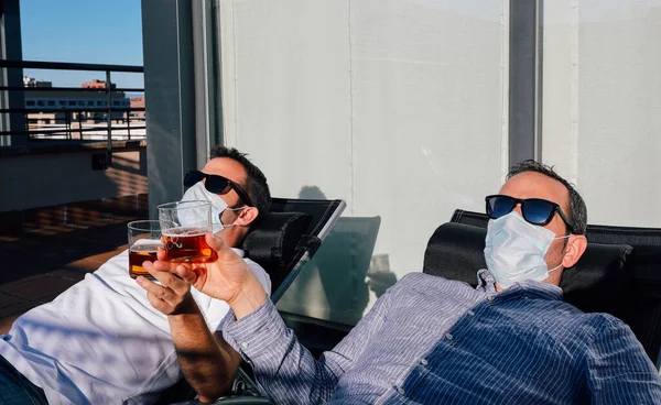 Portrait of two men with faces covered by mask and sunglasses enjoying the summer lying on the roof of a building and toasting with beer