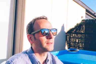 Güneşli bir günde güneş gözlüklü, kendine güvenen yakışıklı bir adamın yakın görüntüsü. Koca, güneş gözlüklerine yansıyan karısı ve kızıyla konuşuyor.