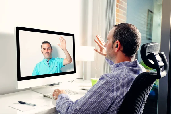 Bilgisayarın önündeki genç iş adamı iş arkadaşıyla görüntülü görüşme yapıyor. Ev ofisinden gelen görüntülü arama için el sallıyor. Arkadaşlar birbirini selamlıyor.