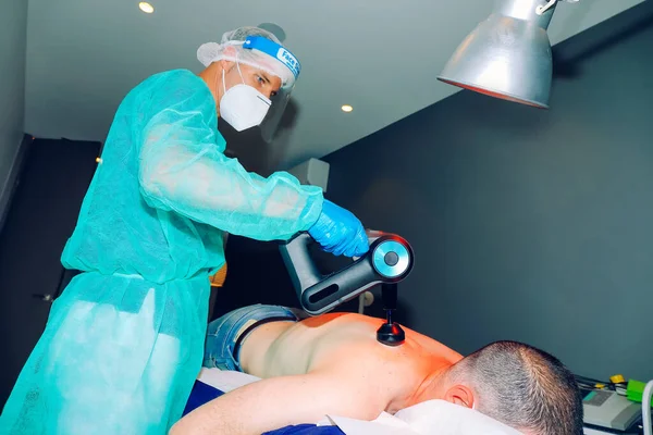 Fizyoterapist hastanın sırtındaki kasılmaları gevşetmek için perküsyon masaj silahı kullanıyor. Koronavirüsü önlemek için yüz kalkanı ve maskesi