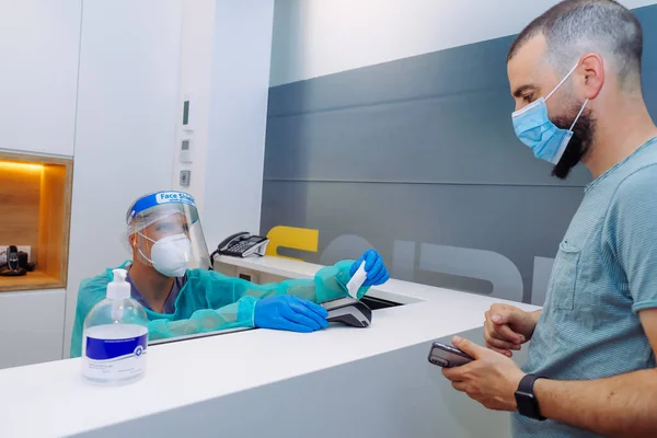 Healthcare worker and client at the reception of a clinic wearing face shield, mask and gloves as precaution against coronacirusvirus. Man receiving ticket after paying