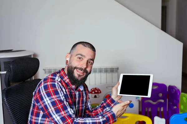 Young smiling man talking to camera and pointing to tablet with blank screen. Work from home. Conference video call and business presentation concept