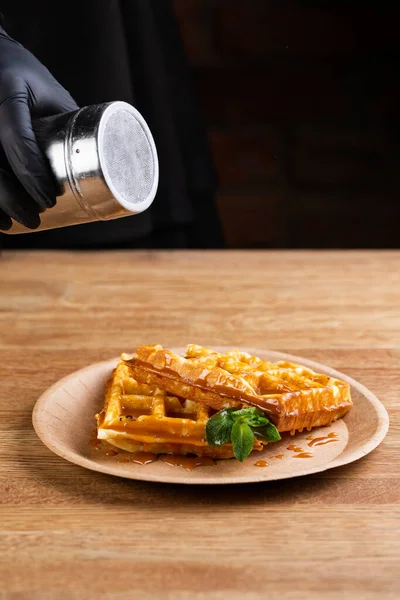 Viennese waffles on a disposable plate with caramel syrup, icing sugar and mint. chef decorates dessert with powdered sugar