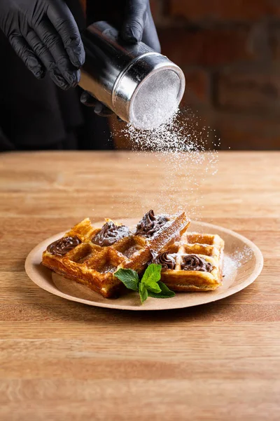 Viennese waffles on a disposable plate with peanut butter, icing sugar and mint. chef decorates dessert with powdered sugar