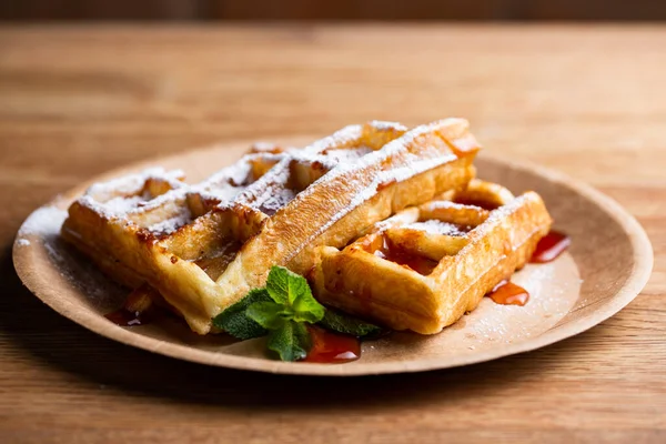 Viennese waffles on a disposable plate with strawberry jam, icing sugar and mint. cook decorates dessert with icing sugar