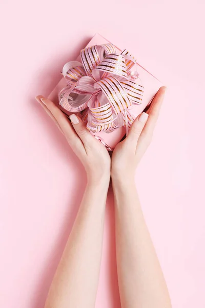 Manos de mujer sosteniendo la caja presente con lazo rosa sobre fondo rosa pastel. Estilo laico plano . — Foto de Stock