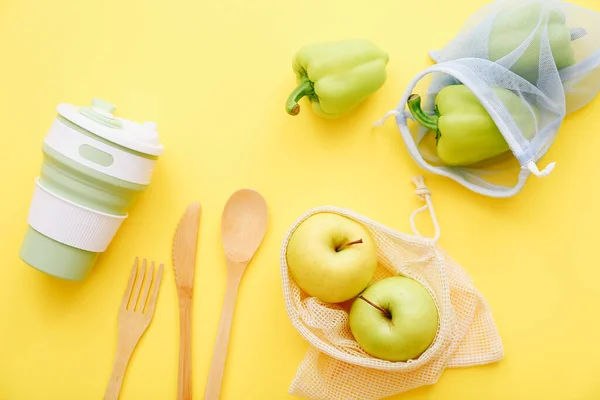 Сітка сумки з яблуками та перцем з багаторазовою чашкою та дерев'яними столовими приборами на жовтому тлі. Вид зверху. Екологічно дружня концепція — стокове фото