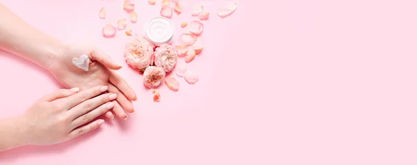 Concepto de cuidado de la piel. Manos femeninas con flores de crema y rosas sobre fondo rosa. Copiar espacio — Foto de Stock