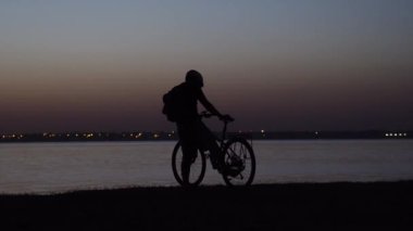 Kasklı bisikletçi oturur ve uzaklaşır. Gün batımına karşı siluet.