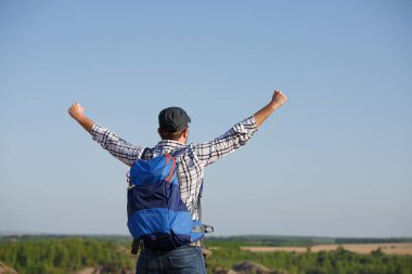 Photo back of tourist man with backpack ,hands up on hill in background of mountain expanses, blue sky clipart