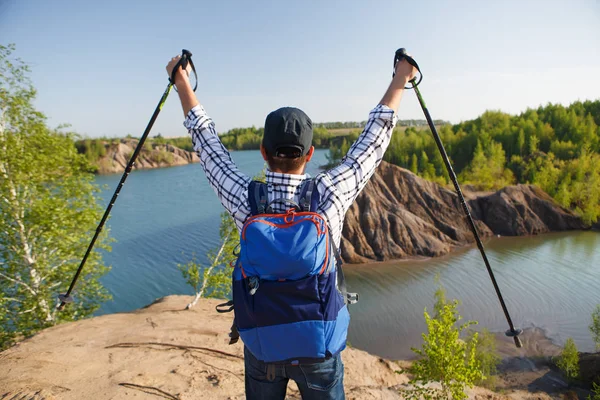 Photo from back of tourist man with walking sticks with his hands up on mountain hill near lake — Stock Photo, Image