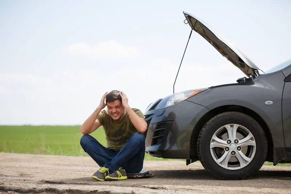 Image of young man sitting next to broken car with open hood — Stock Photo, Image