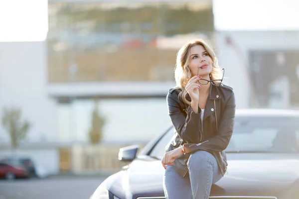 Фото молодой женщины в солнечных очках, смотрящей в сторону, сидящей на капоте черной машины — стоковое фото