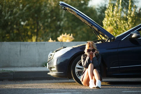 Photo of young woman sitting near broken car with open hood