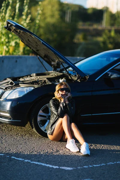 Obrázek blondýnka sedí na asfaltu vedle rozbité auto s otevřenou kapotou — Stock fotografie