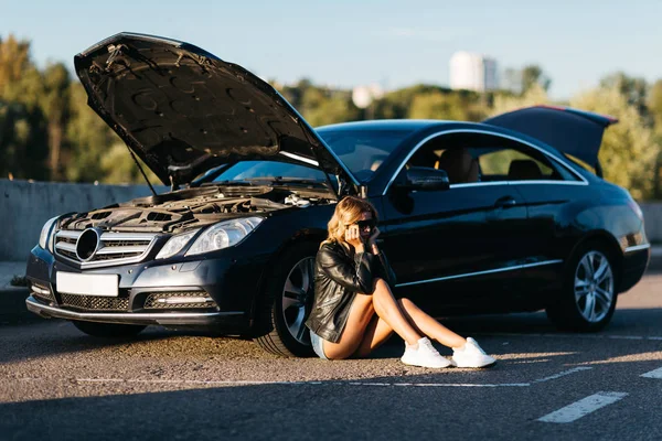 Imagem de mulher chateada sentada no asfalto ao lado de carro quebrado com capuz aberto — Fotografia de Stock