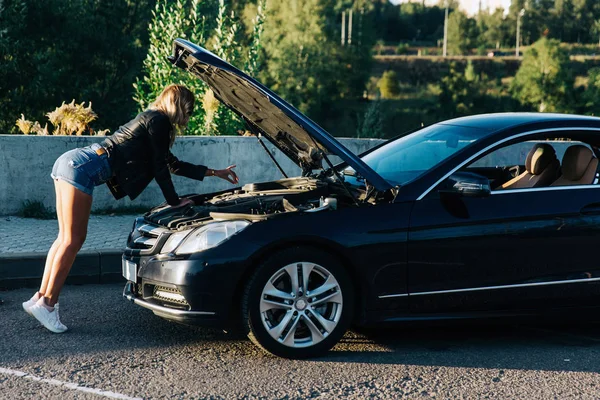 Изображение на стороне молодой блондинки, стоящей перед черной машиной с открытым капотом — стоковое фото