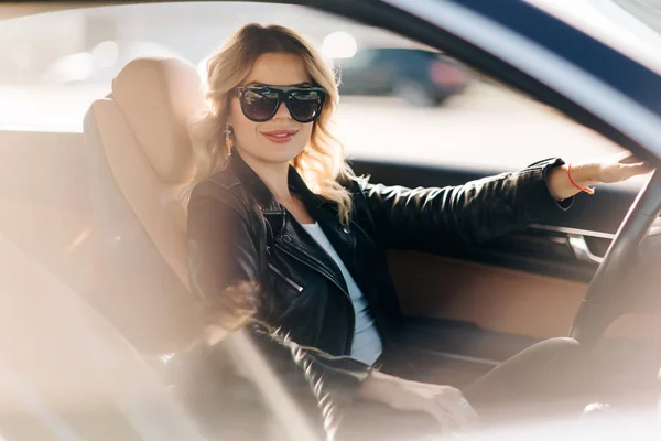 Foto de loira de cabelos longos com cabelo encaracolado em óculos pretos sentados em carro preto . — Fotografia de Stock
