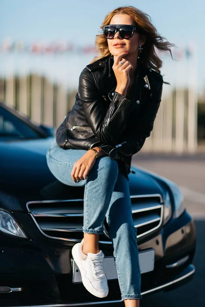 Portrét mladé blond ženy v sedící na kapotě auta černé sluneční brýle — Stock fotografie