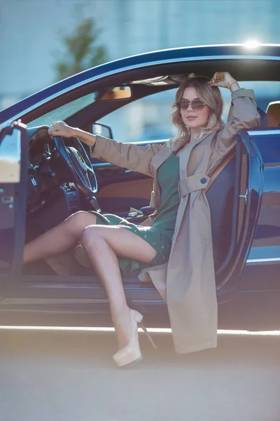 Фото женщины в очках и длинном платье и туфлях на высоких каблуках, сидящей в машине с открытой дверью — стоковое фото