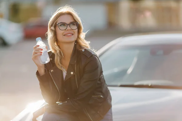 Изображение блондинки в очках с бутылкой воды в руках, сидящей на капоте автомобиля — стоковое фото