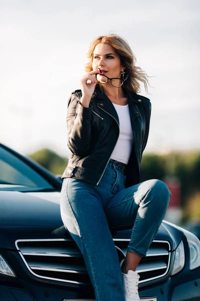 Porträt einer jungen Blondine mit Brille in den Händen, die auf der Motorhaube eines schwarzen Autos sitzt — Stockfoto