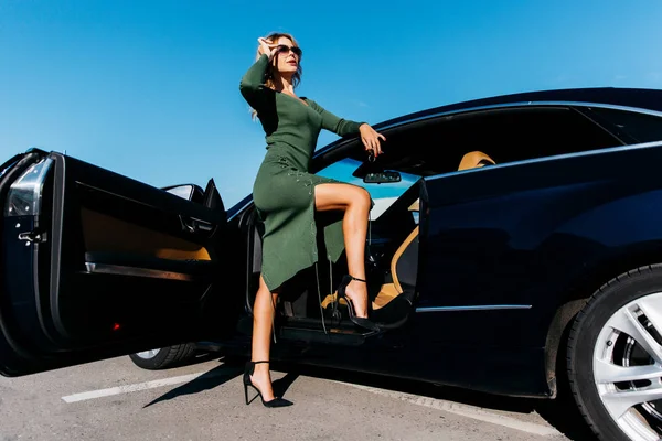 Foto di bionda sexy con chiavi in occhiali da sole in abito lungo in piedi vicino auto nera con porta aperta — Foto Stock