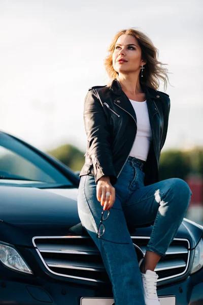 Elinde siyah bir araba kaputa oturan gözlüklü genç sarışın fotoğrafı