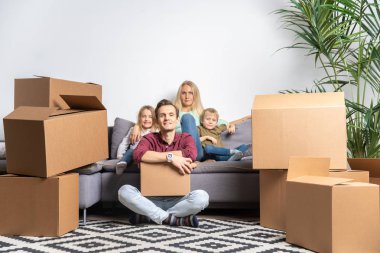 Karton kutular arasında gri kanepede oturan kız ve erkek ile kadın ve zemin üzerinde oturan adam fotoğrafı