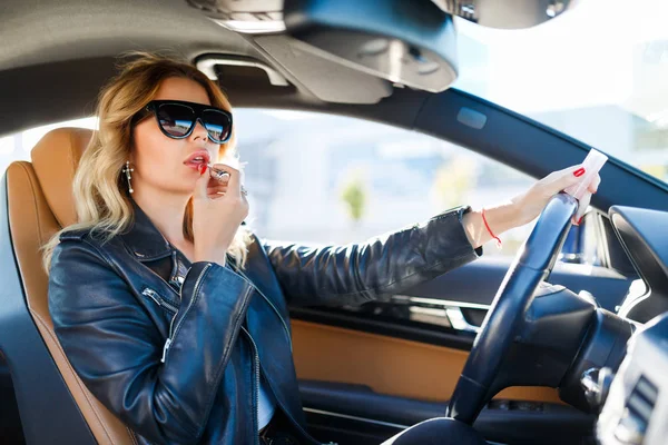 Gambar si pirang muda berkacamata hitam mengecat bibir yang duduk di mobil — Stok Foto