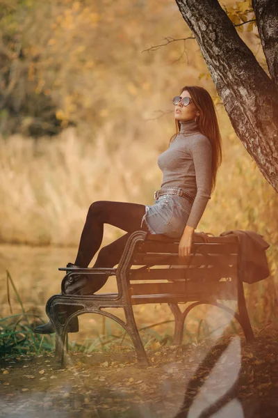 Изображение женщины в солнцезащитных очках, сидящей на скамейке в осеннем парке — стоковое фото