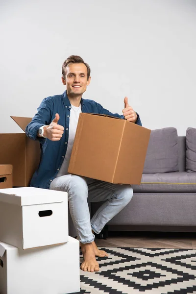 Foto de la morena feliz sentada en el suelo con cajas de cartón en el fondo del sofá gris —  Fotos de Stock