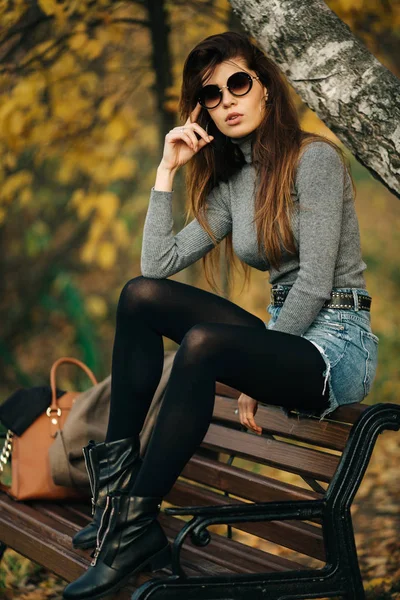 Foto av brunett i solglasögon sitter på bänken i höst park — Stockfoto