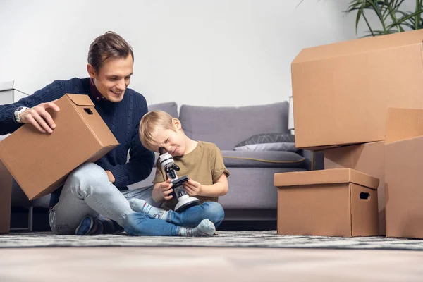 Photo of happy man and small boy with microscope sitting on floor among cardboard boxes — Stock Photo, Image