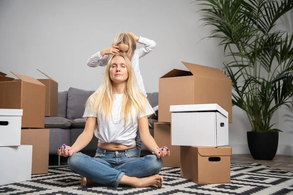 Photo of calm woman sitting on floor among cardboard boxes and girl jumping on sofa