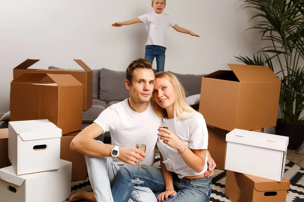 Фото молодой пары с бокалами для вина, сидящей на полу, и сына на диване среди картонных коробок — стоковое фото