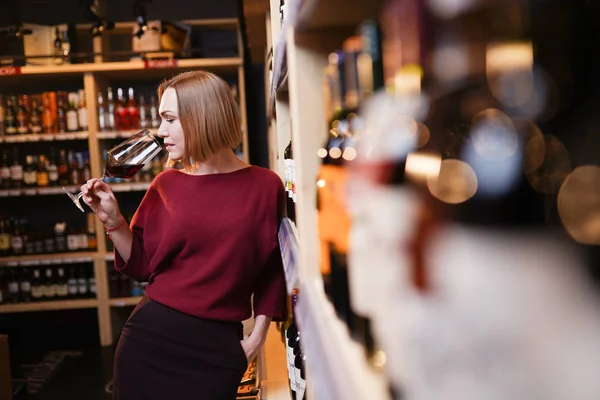 Immagine di giovane donna con bicchiere di vino in negozio sullo sfondo di scaffali — Foto Stock