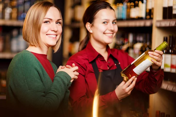 Foto di due donne sorridenti con bottiglia di vino in serbo sullo sfondo di scaffali — Foto Stock