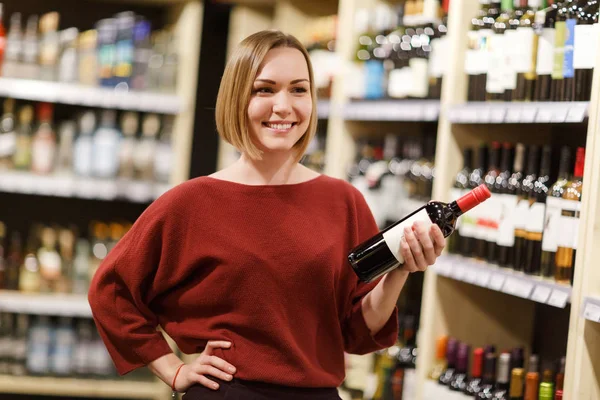 Image de femme heureuse avec bouteille de vin sur fond flou d'étagères avec des bouteilles — Photo