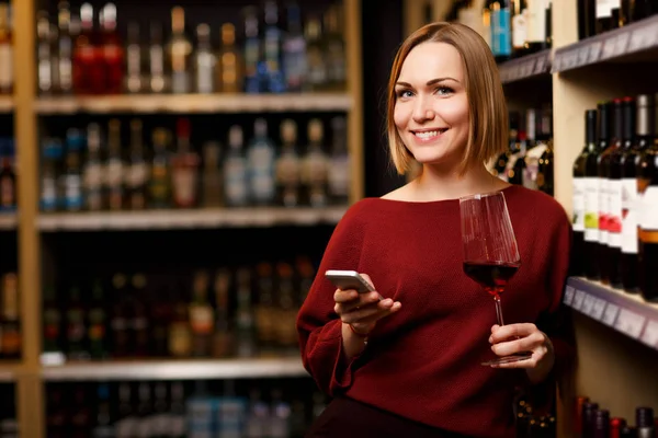Image de blonde heureuse avec téléphone et verre dans les mains au magasin avec du vin — Photo