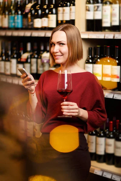 Foto de jovem loira com vidro nas mãos na loja com vinho — Fotografia de Stock