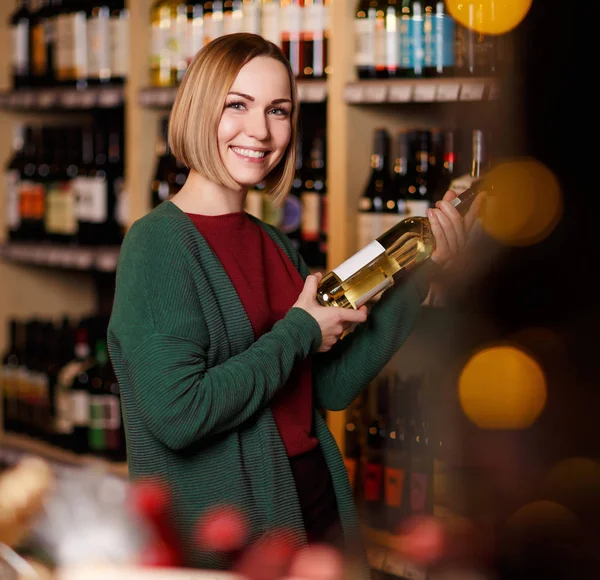 Immagine di donna felice con bottiglia in mano su sfondo sfocato di scaffali con bottiglie di vino — Foto Stock