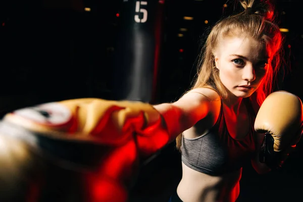 Blonde sportswoman in boxing gloves hits bag in training.