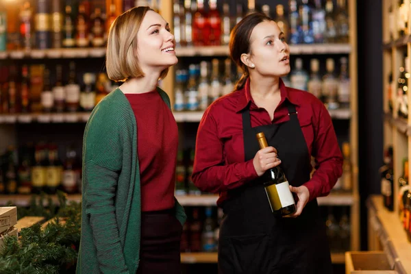 Foto di due giovani donne su sfondo sfocato di scaffali con bottiglie di vino — Foto Stock