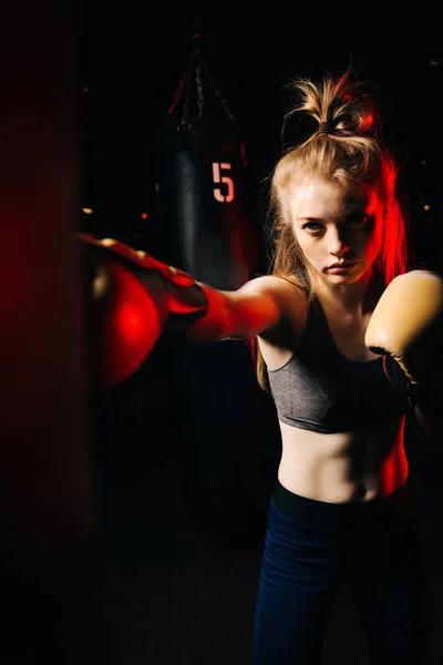 Blonde sportswoman in boxing gloves hits bag in training, red light.
