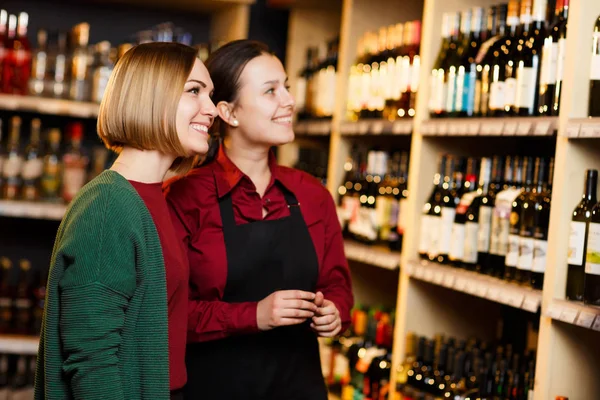 Imagem de duas mulheres sorridentes no fundo borrado de prateleiras com garrafas de vinho — Fotografia de Stock