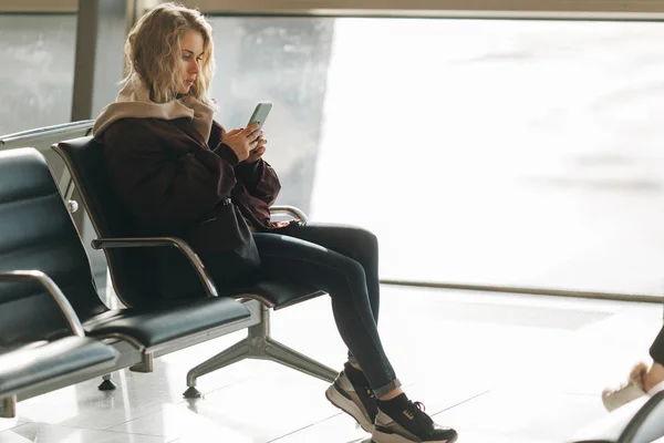 Кудрявая блондинка с телефоном в руках, сидящая в комнате ожидания  . — стоковое фото