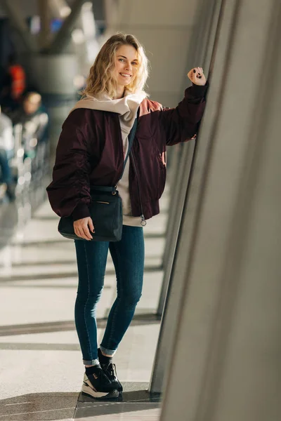 Feliz rubia rizada mirando a la cámara se para en la sala de espera . —  Fotos de Stock