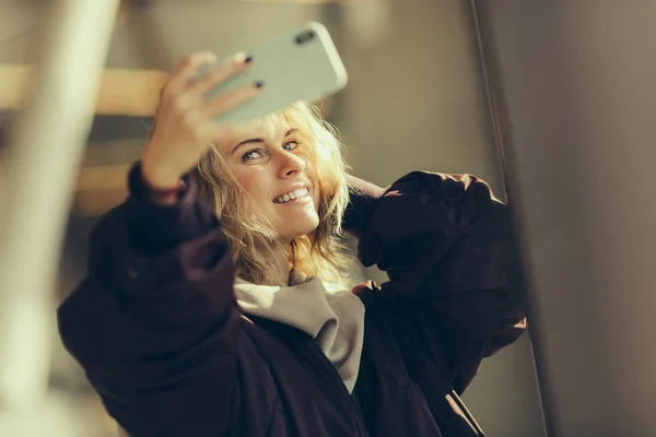 Junge glückliche Blondine in Jacke macht Selfie vor Fensterhintergrund — Stockfoto